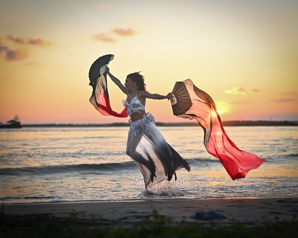 Dancer twirls flowing red and black fabric on beach at sunset creating dramatic silhouette against golden sky.