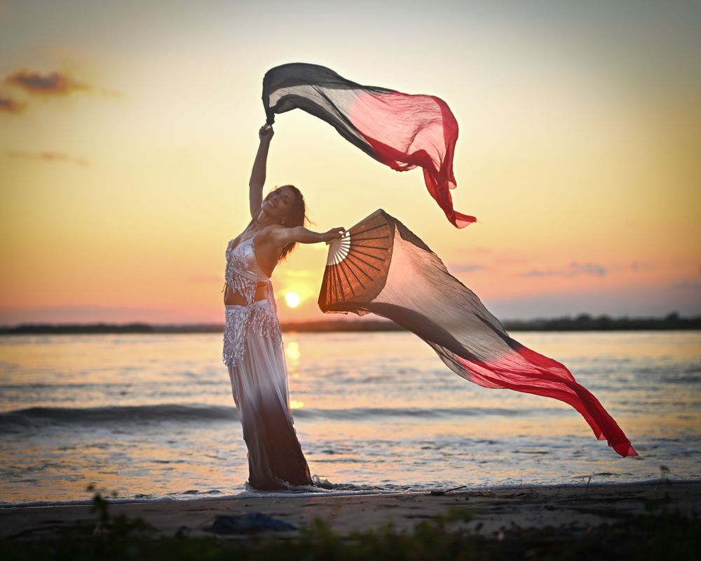 Figure dancing with flowing red and black fabric on beach during golden hour.