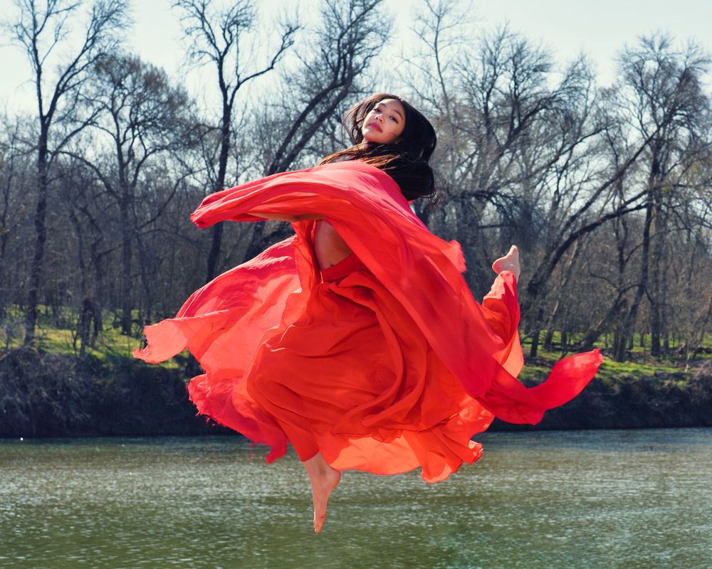 Dancer leaps gracefully in flowing red dress by riverside with bare trees in background.