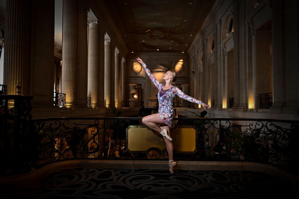 Ballet dancer performs graceful leap in grand columned hallway with ornate railings.