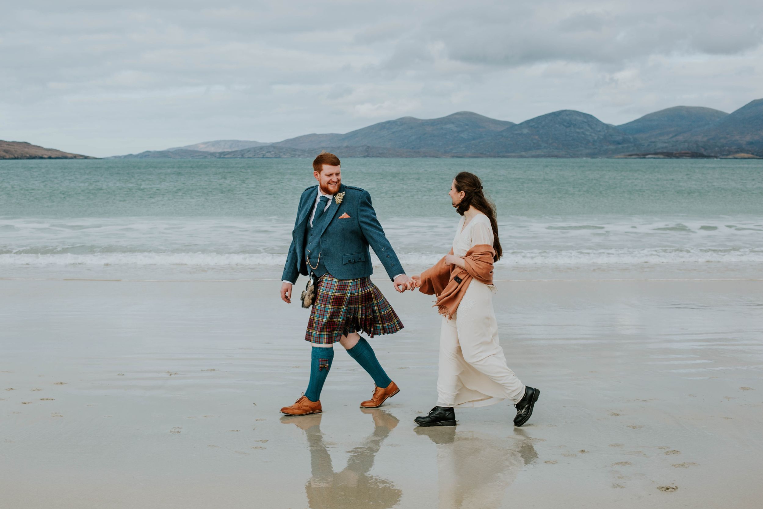 Couple playfully walking along Luskentyre beach with mountains in background, one wearing traditional Scottish kilt.