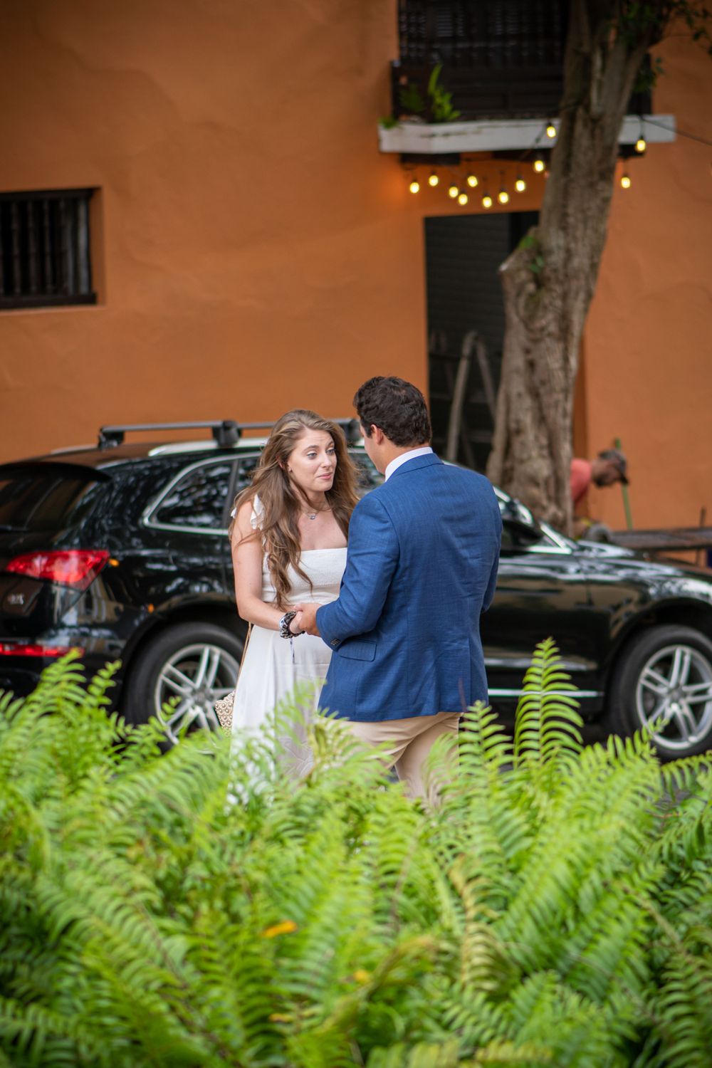 Couple sharing a romantic moment in front of an orange wall with ferns in foreground.