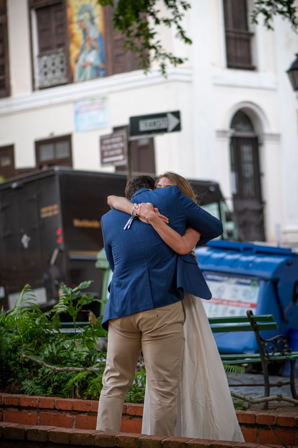 Couple embracing in front of urban building with plants in foreground.
