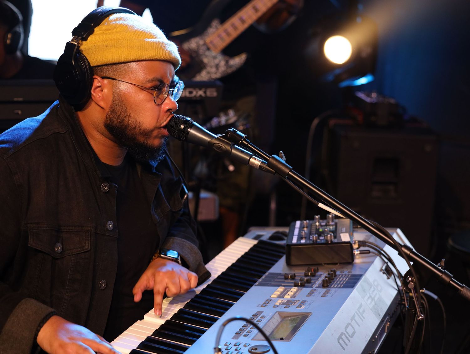 Musician wearing yellow beanie plays keyboard and sings into microphone during live performance on dark stage.