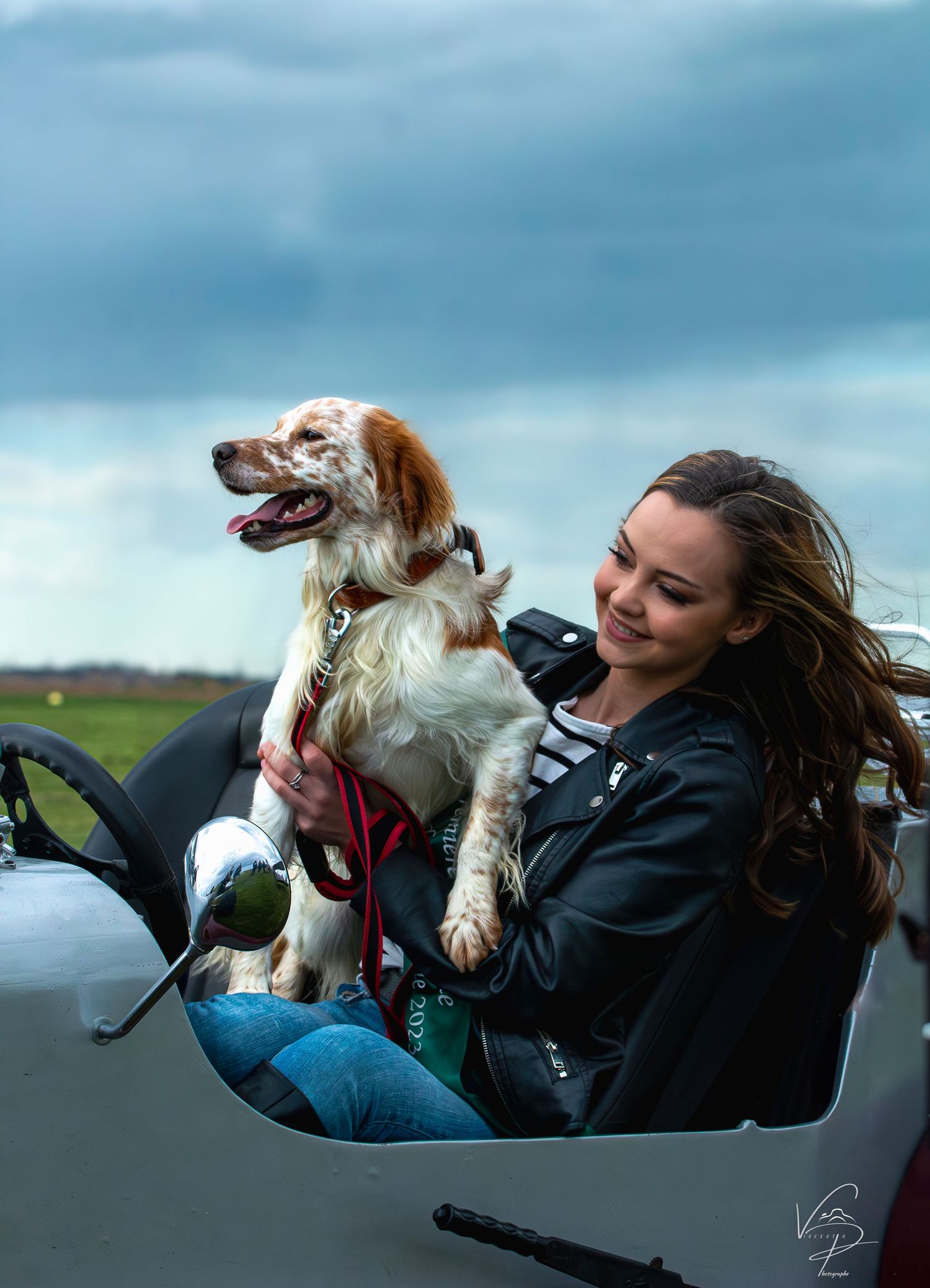A brown and white dog enjoys a playful moment in a car on a cloudy day.