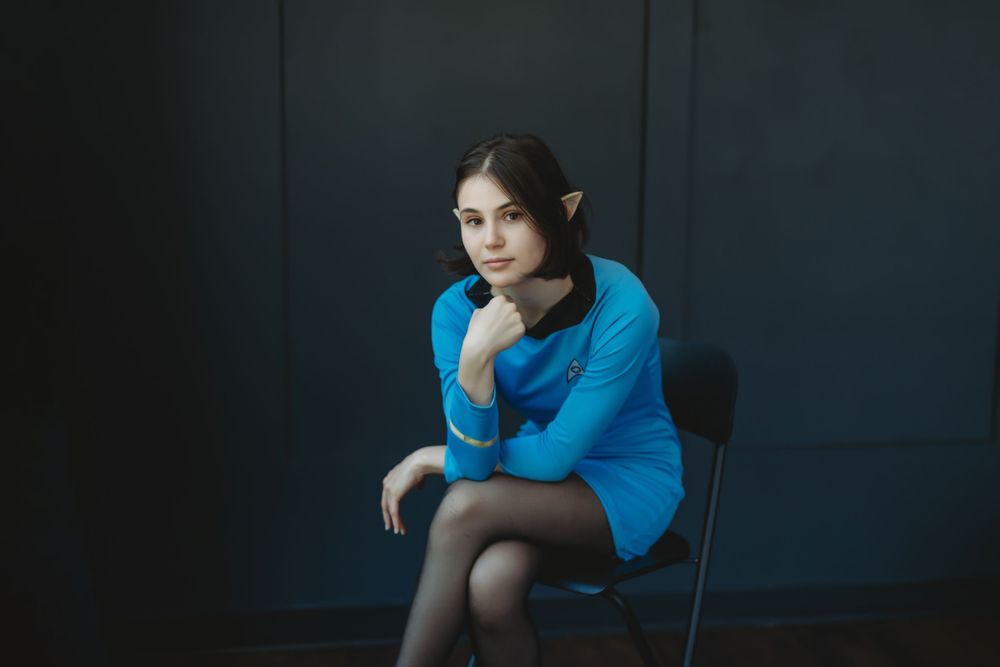 A person in a bright blue sweater sits thoughtfully on a chair against a dark backdrop. Bellevue studio, WA