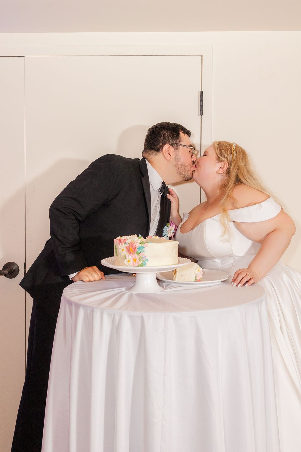 A couple in wedding attire share a kiss at a table with a floral wedding cake on a white tablecloth.