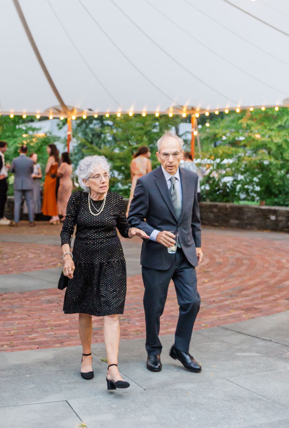 Elderly couple playfully dancing together on a brick patio with flowers and greenery in the background.