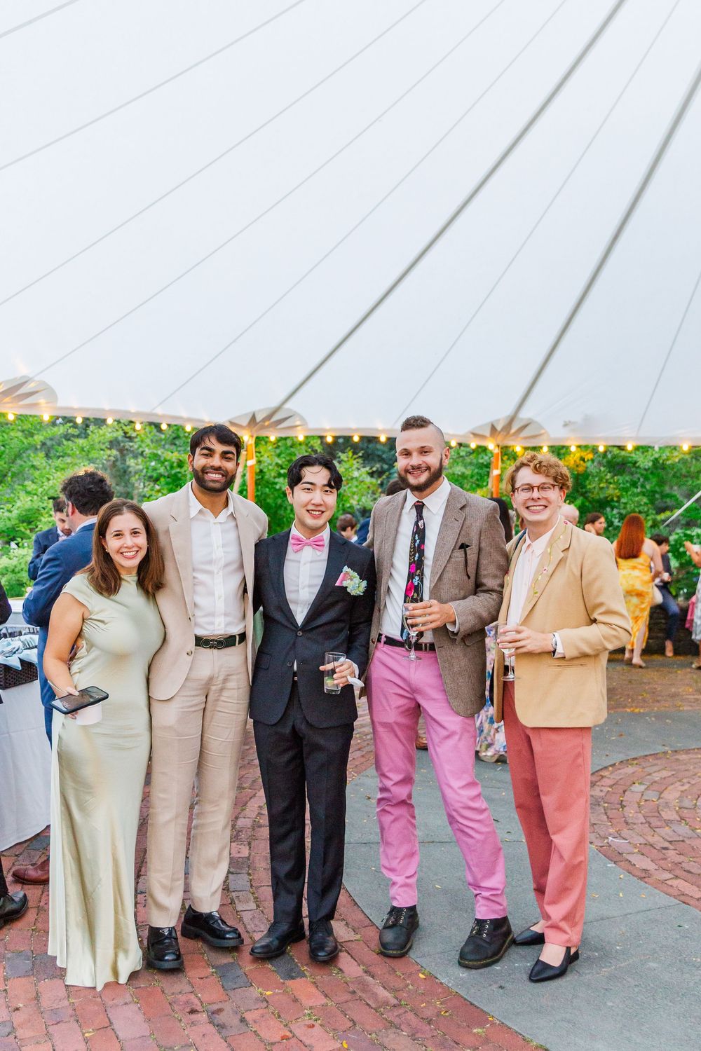 A group of wedding guests wearing pastel and neutral colored formal attire pose together on a brick pathway.