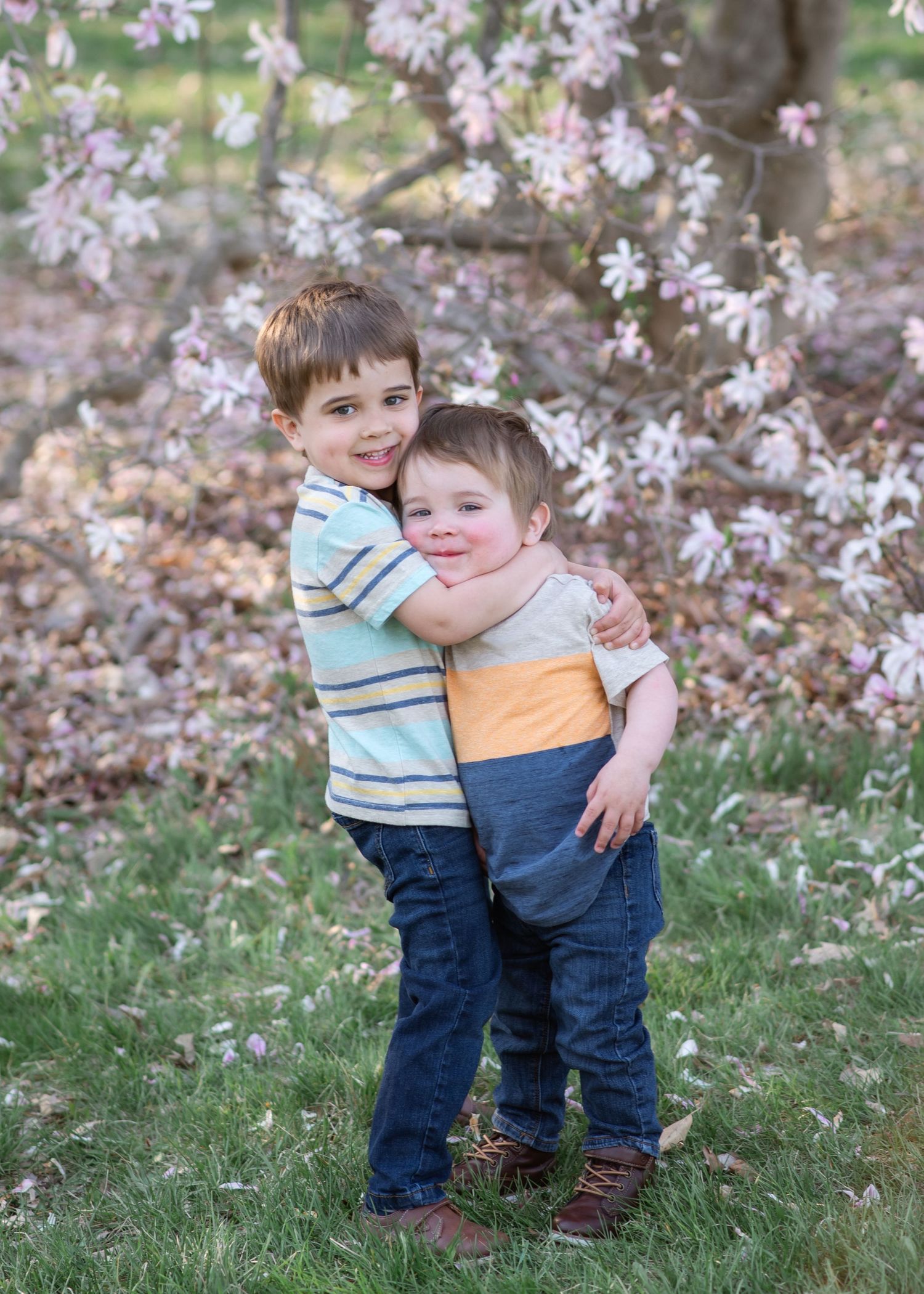 Two young children embrace while standing in front of blooming cherry blossom trees on a spring day.
