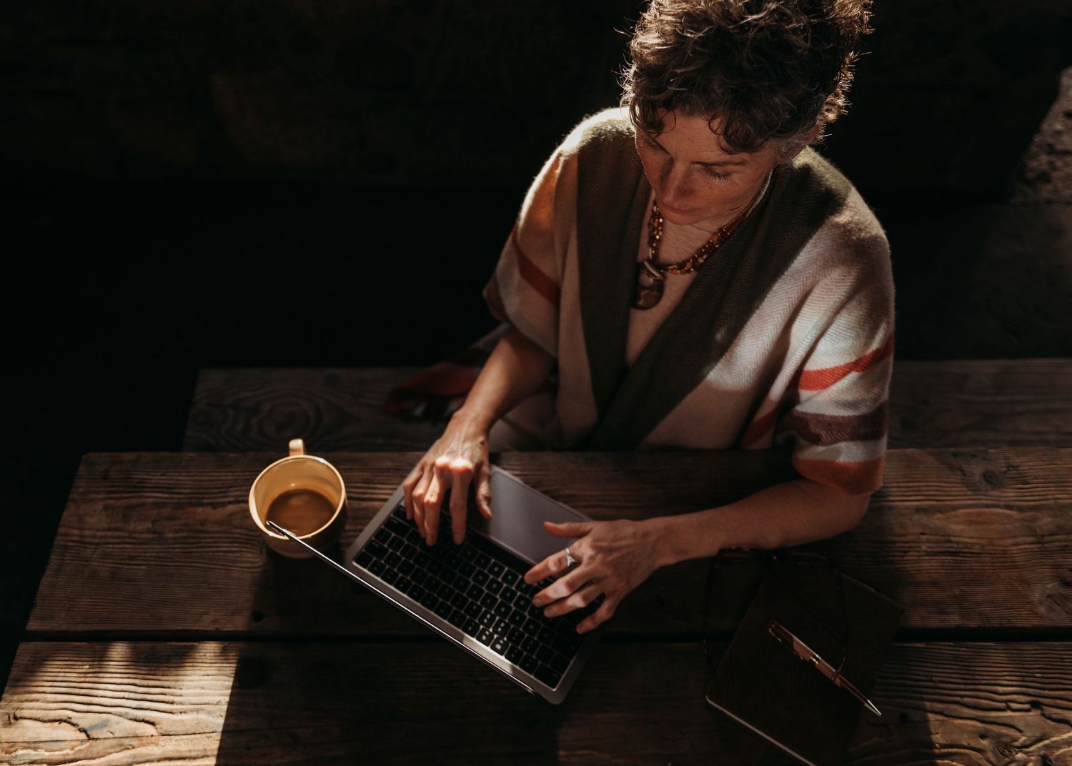 Person in moody lighting working at wooden table with laptop and coffee cup in rustic setting.