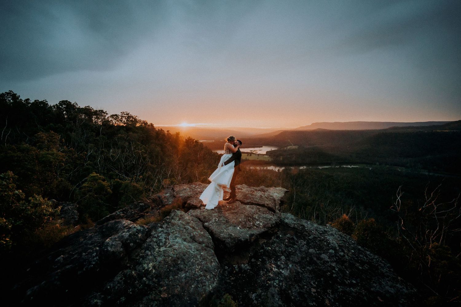 Dramatic clifftop wedding portrait at sunset overlooking mountain valley.