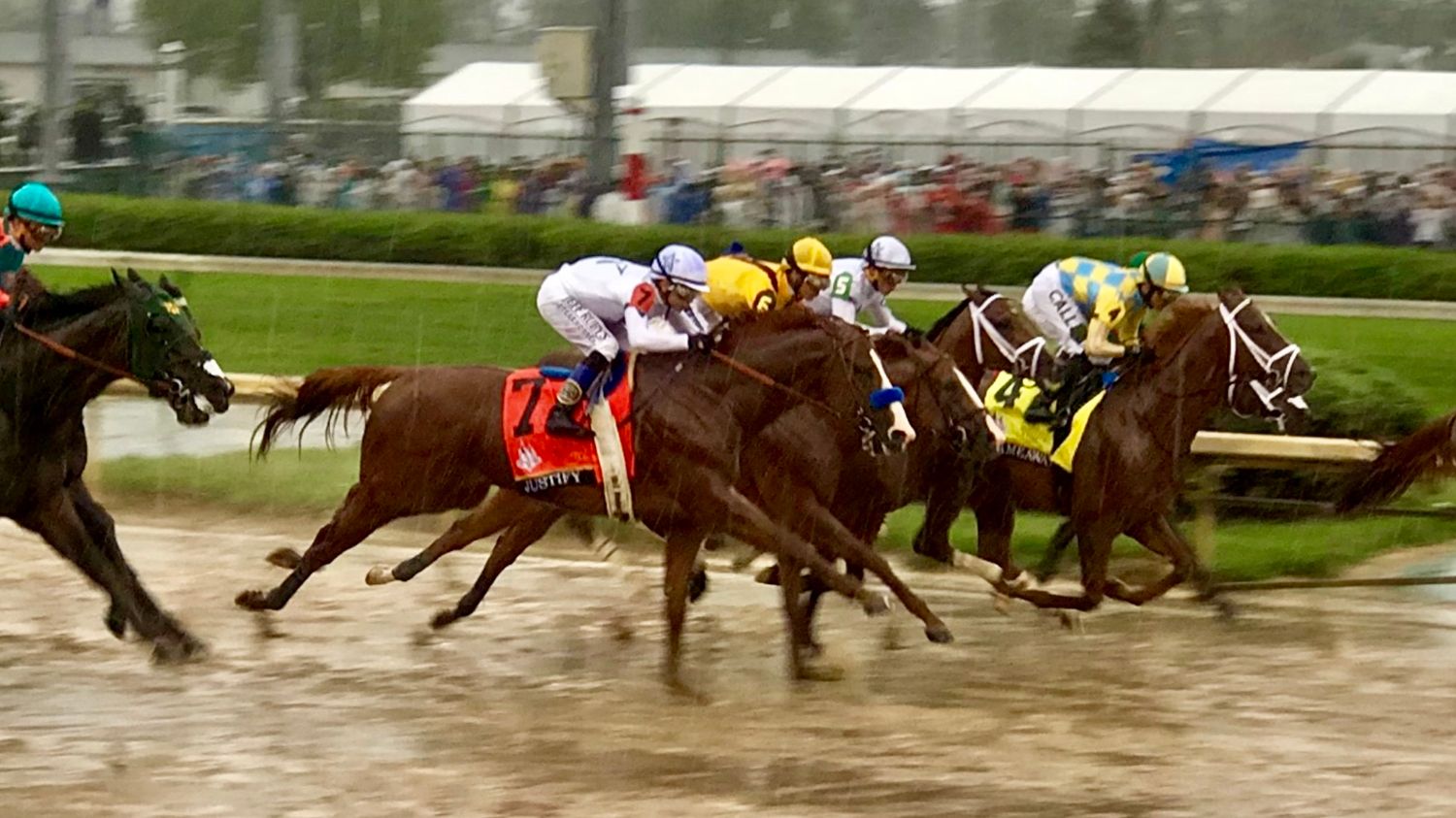 Horse racing action at a racetrack with multiple thoroughbreds competing in close formation on a muddy track.