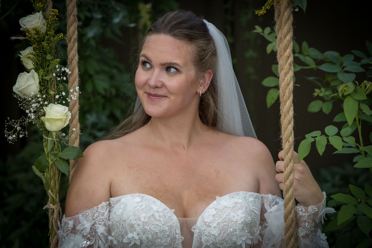 Close-up of smiling bride in off-shoulder lace wedding dress sitting on a rope swing in a garden.