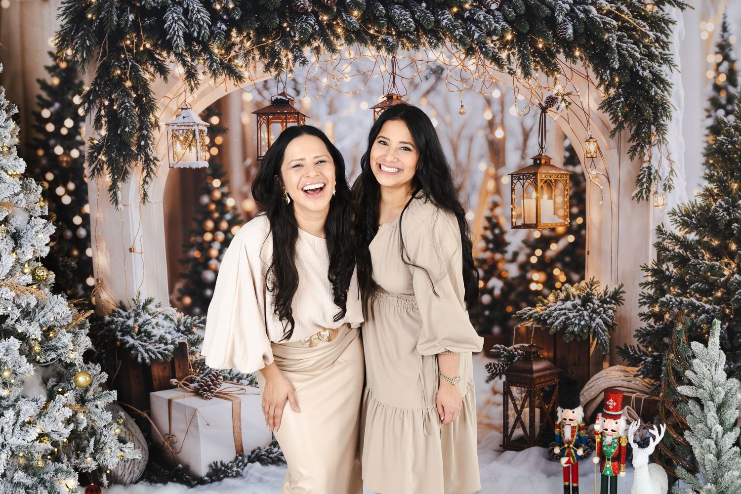 Two women in neutral beige dresses pose together against winter holiday backdrop with lanterns.