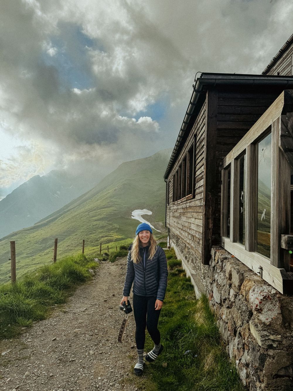 Stone building with windows nestled against a dramatic mountain backdrop under cloudy skies.