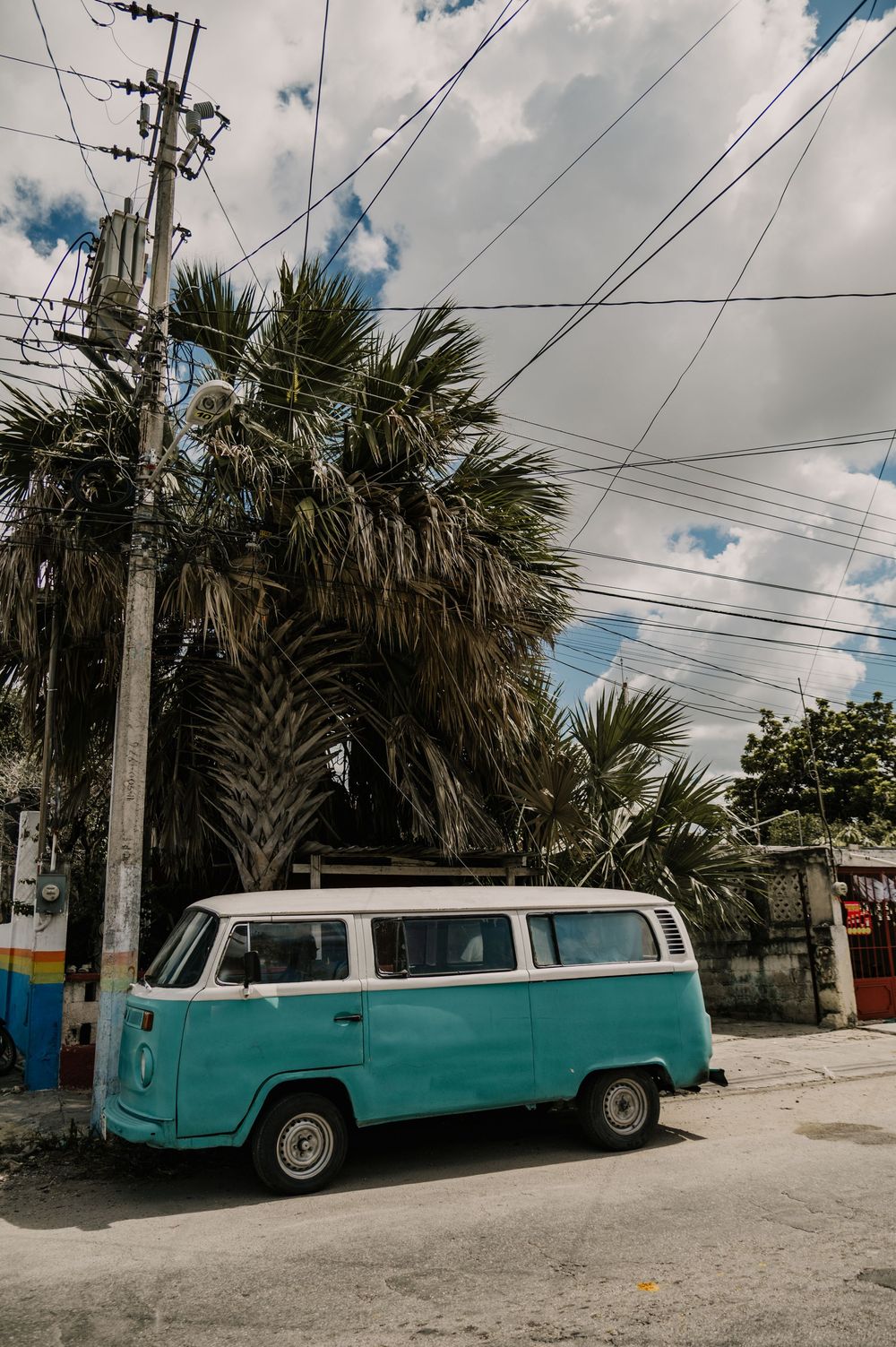 A vintage turquoise Volkswagen van parked on a street lined with palm trees.