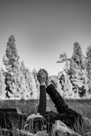 Picture of a couple laying in a field holding hands with trees in the background.