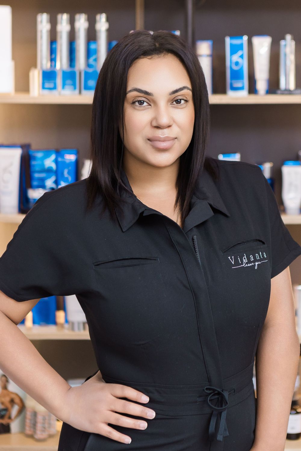 Professional wearing a black collared uniform shirt poses in front of retail shelving displaying blue product packaging.