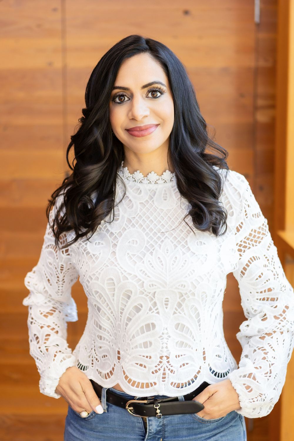 A woman wearing a white lace blouse with bell sleeves and jeans poses against a wooden wall.