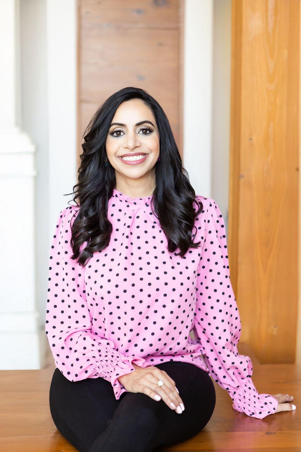 Professional portrait in pink polka dot blouse and black pants sitting on wooden floor with natural lighting.