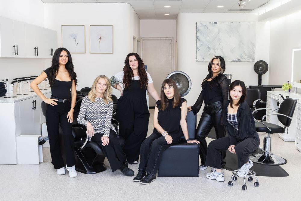 A group in black outfits poses together in various casual positions in a bright white studio space.
