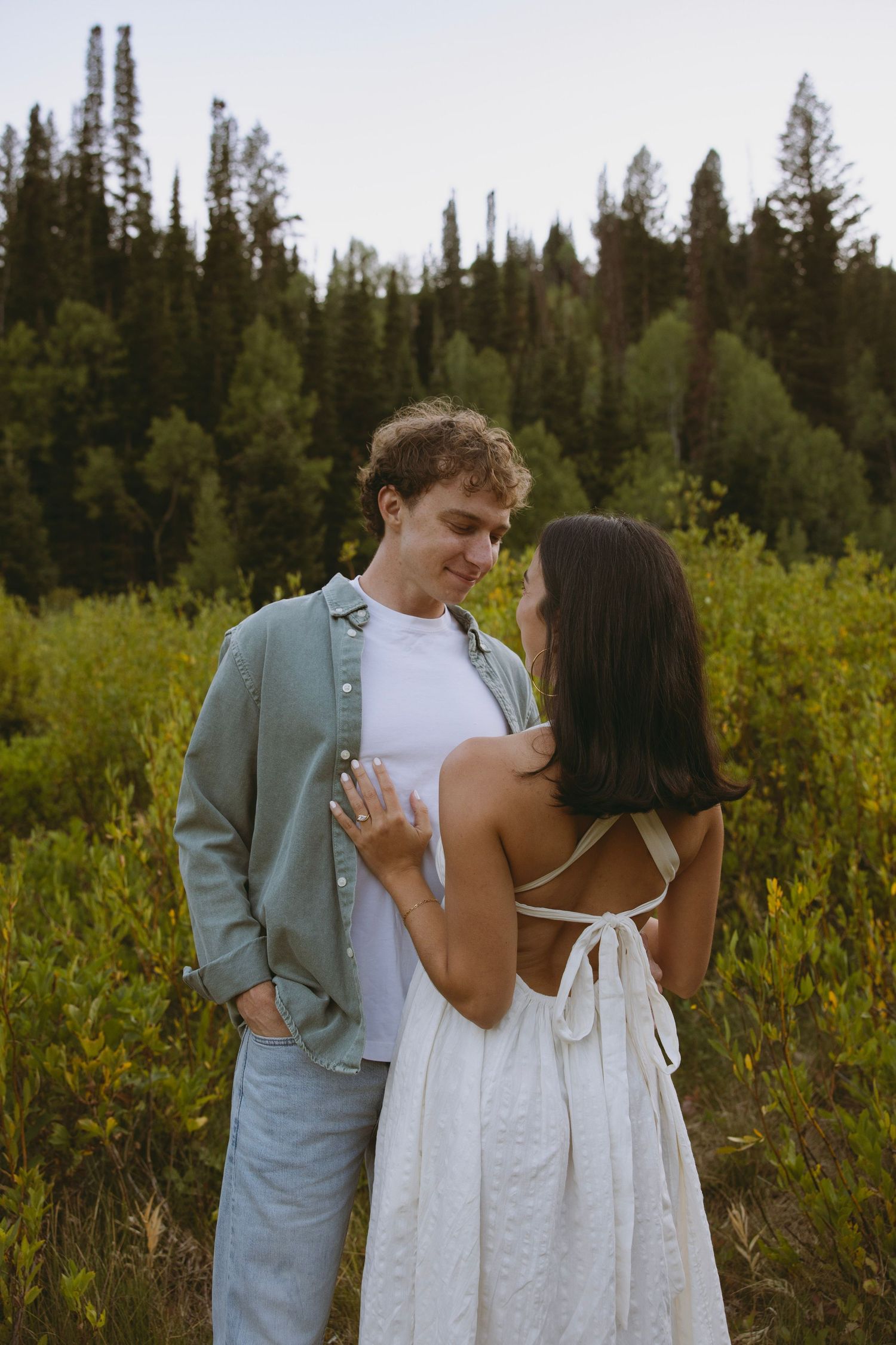A couple shares a romantic moment in a white dress and casual attire against a backdrop of evergreen forest.