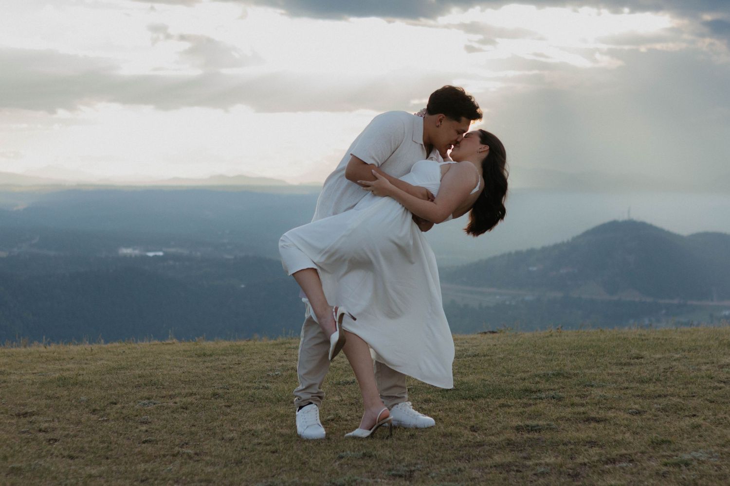 A romantic couple in white outfits share an intimate moment while dancing on a hilltop with mountain views of Colorado in the background.