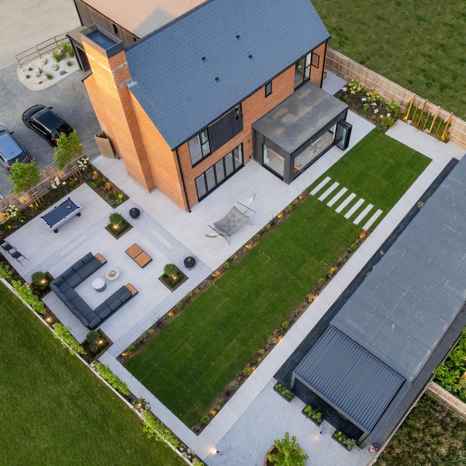 Aerial view of a modern brick house with blue roof featuring a landscaped garden, patio area, and detached garage.
