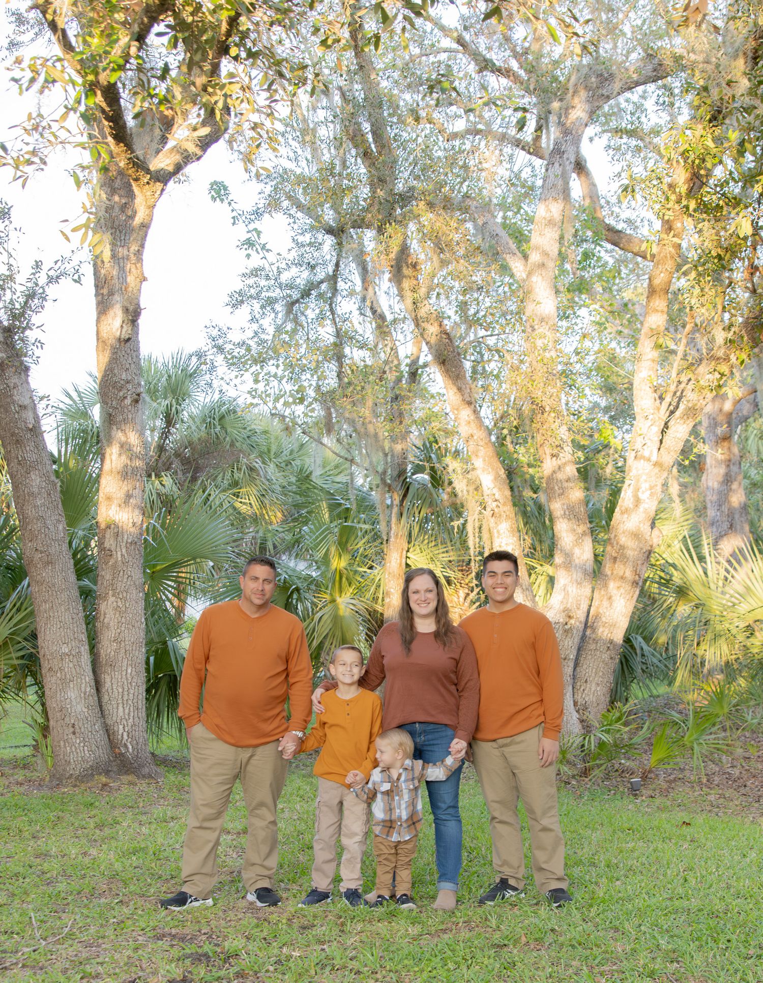 Family of five posing outdoors among tall trees in coordinating orange and tan outfits for a fall portrait session.