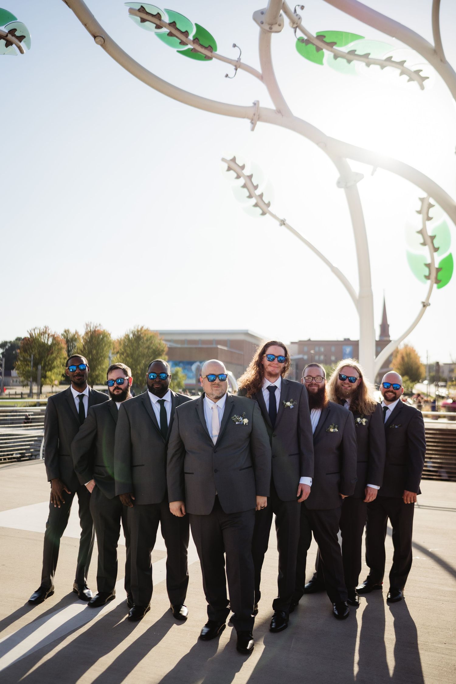 A group of groomsmen in black suits pose together outdoors in a line formation during a wedding photoshoot.