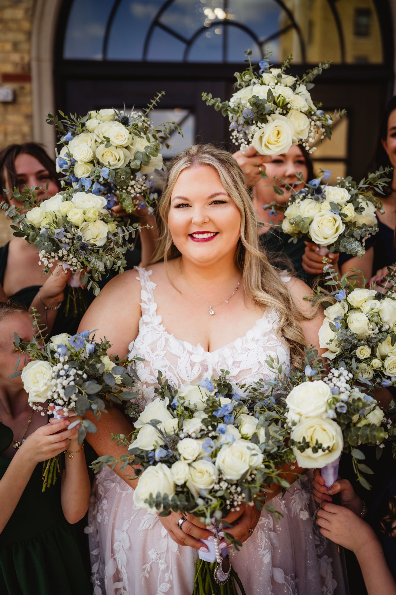 A wedding party poses with elegant white and blue floral bouquets while smiling and laughing together.