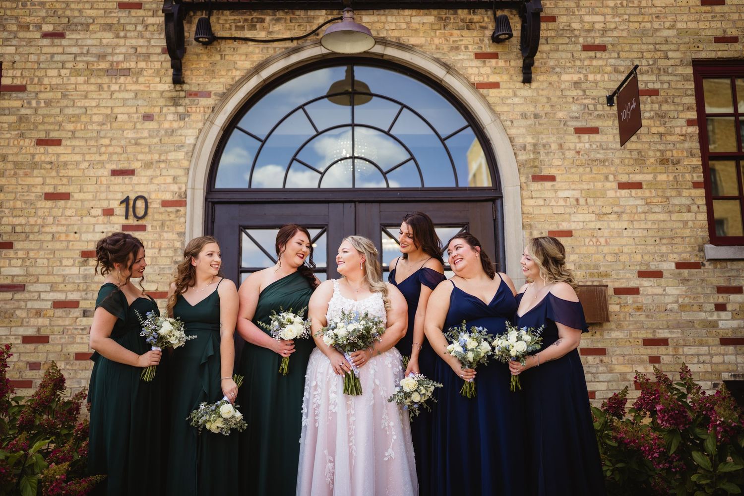 Wedding party stands in front of a brick church wall with an elegant arched window in the background.