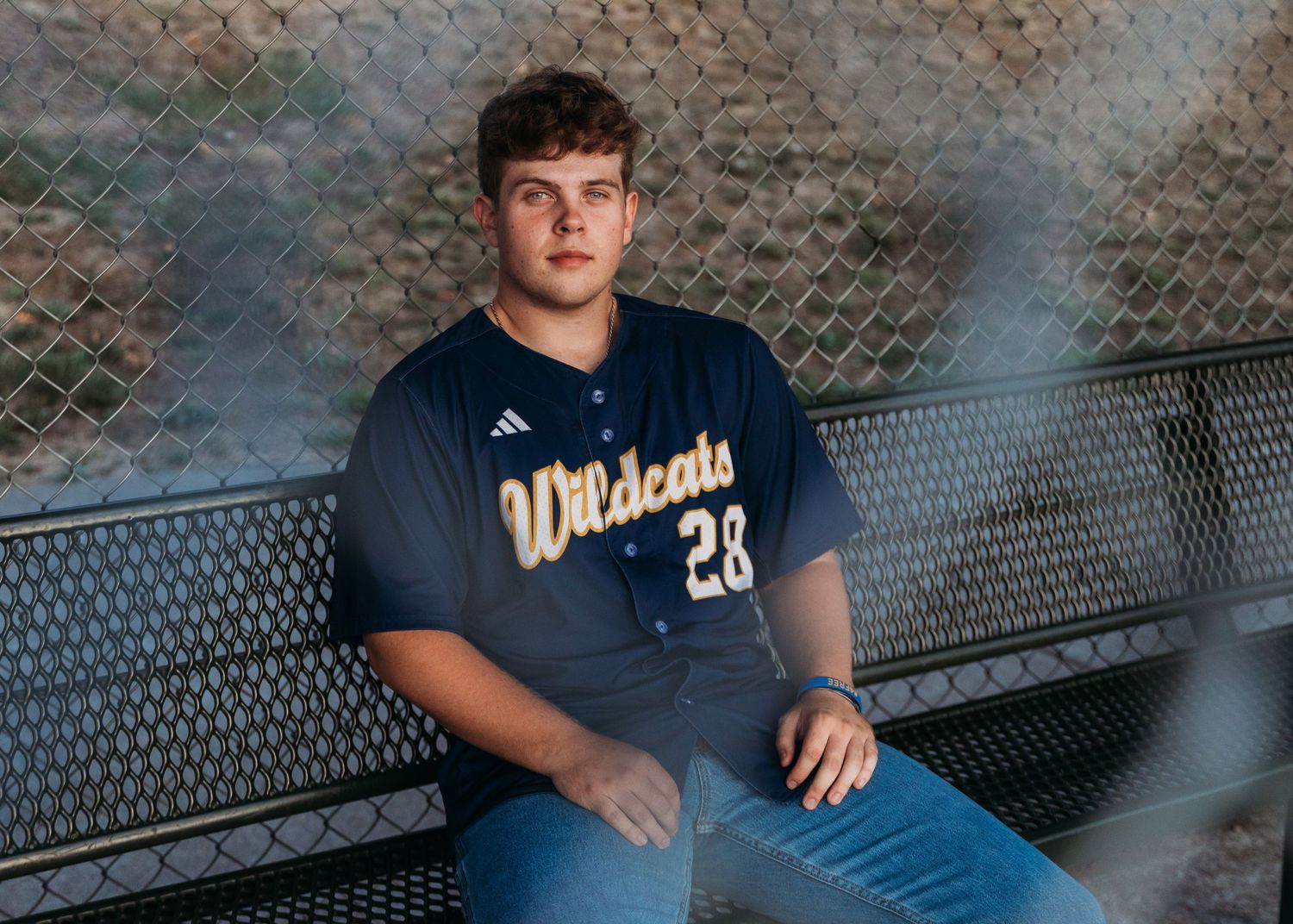 Baseball player wearing a navy blue Wildcats jersey number 28 sits on bench in front of chain link fence.