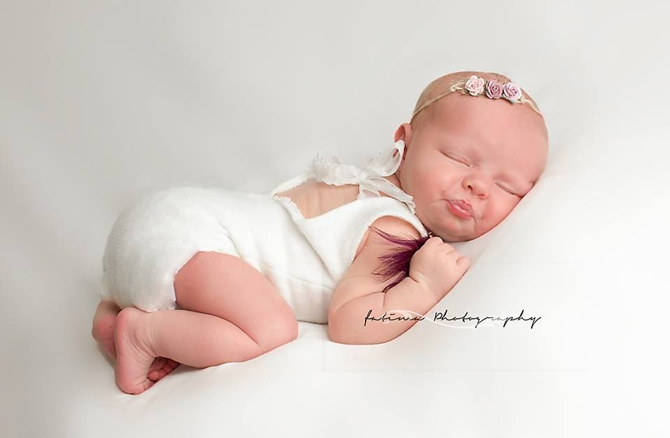 Sleeping newborn baby in white outfit resting on soft white backdrop during professional photo session.