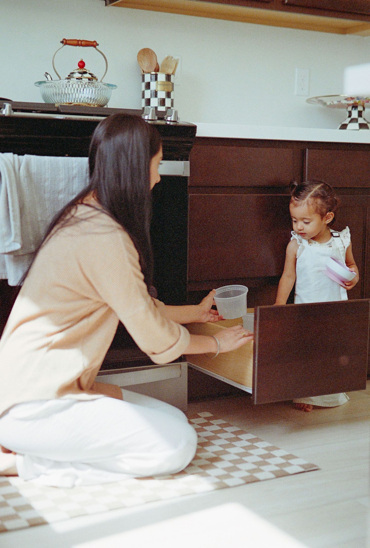 Person in white clothing kneeling on checkered floor while working in a kitchen.