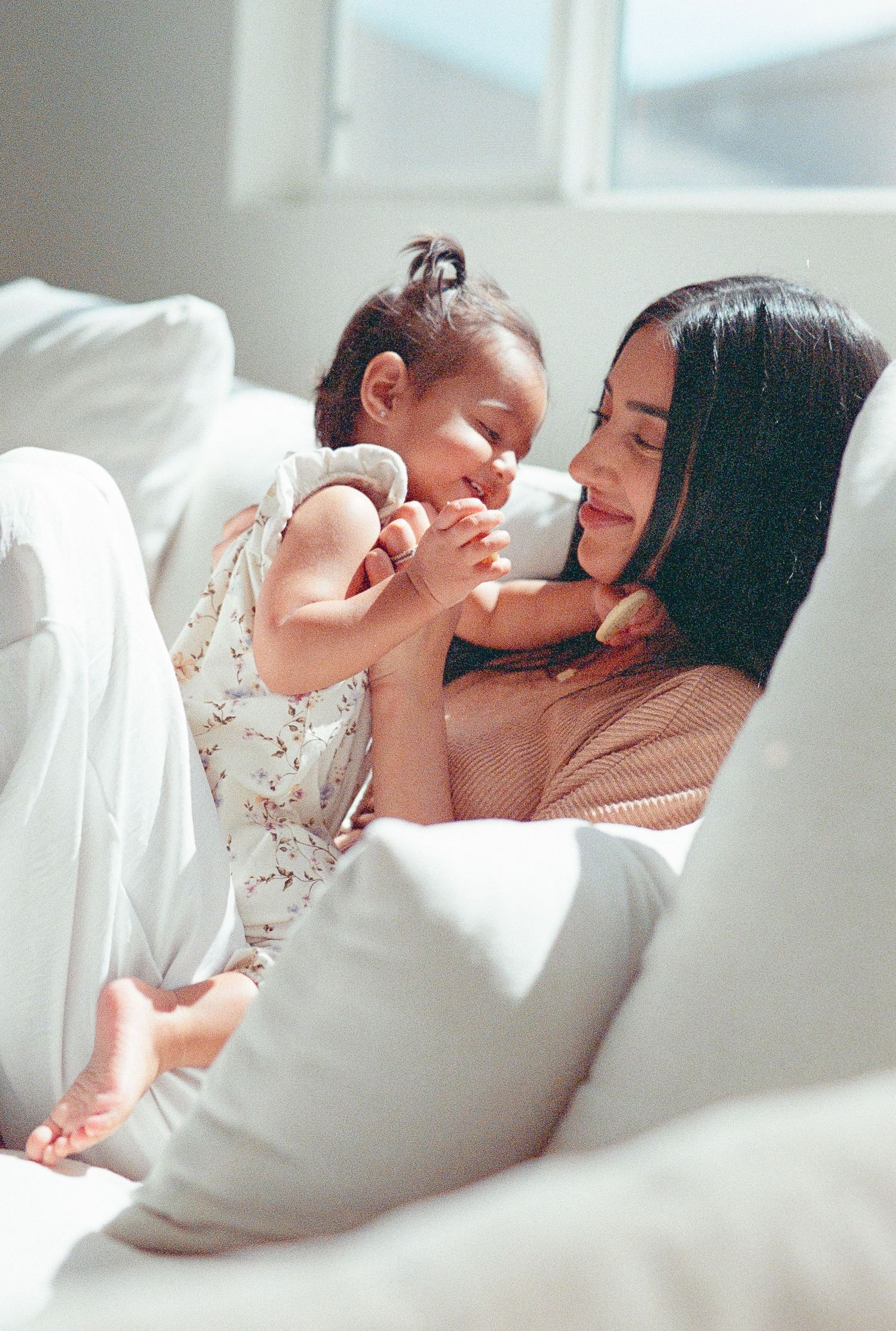 Parent and baby share tender moments together while relaxing on white bedding in soft natural light.