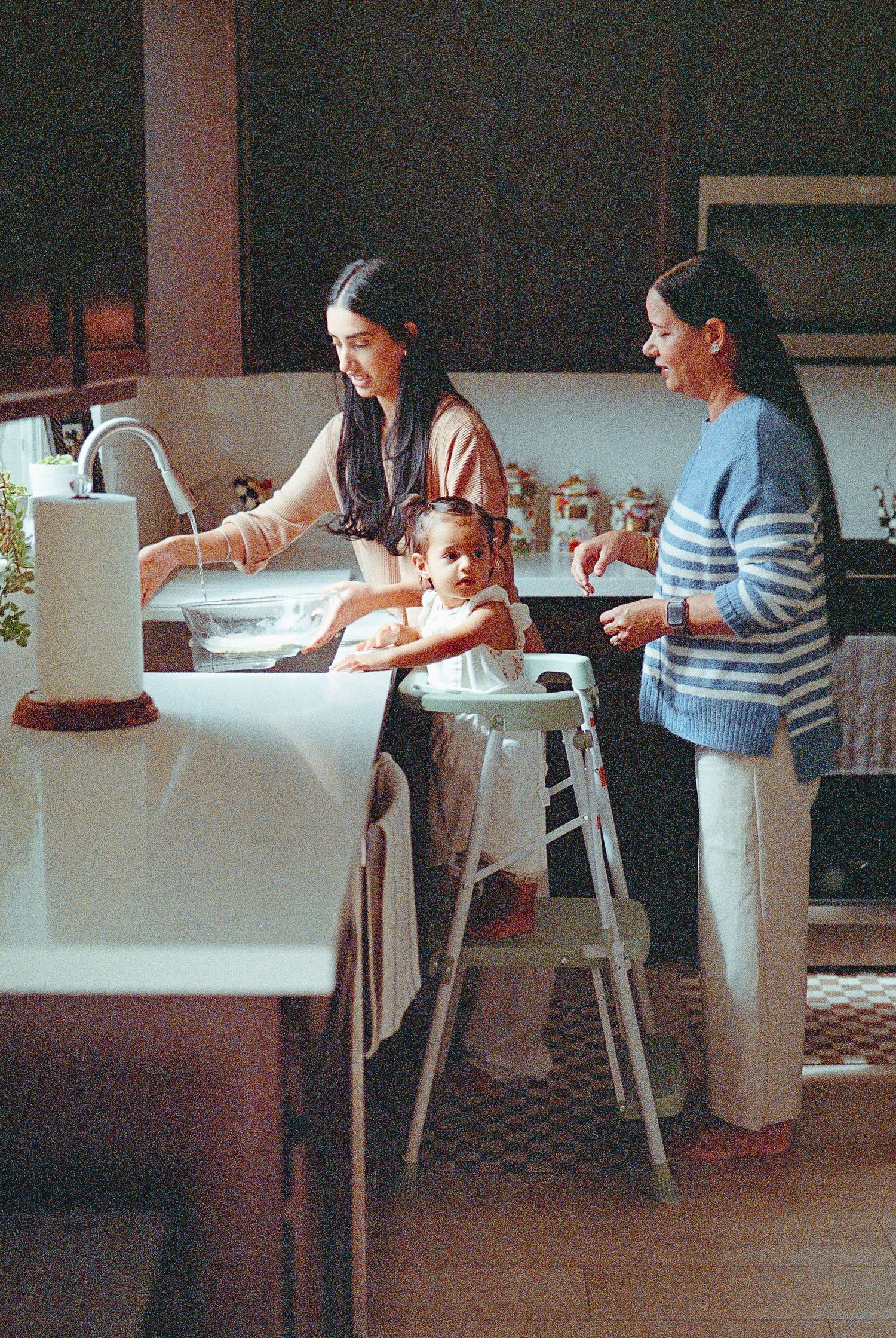 People gather around a kitchen table for a cooking or baking activity in a warm, dimly lit home setting.