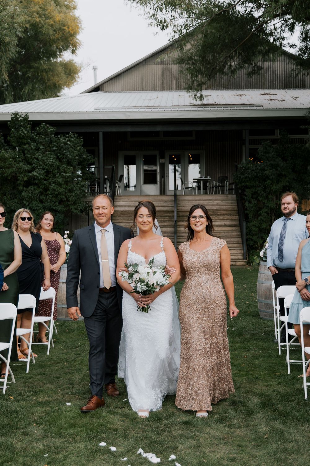 Bride walks down the aisle with parents at an outdoor wedding ceremony.