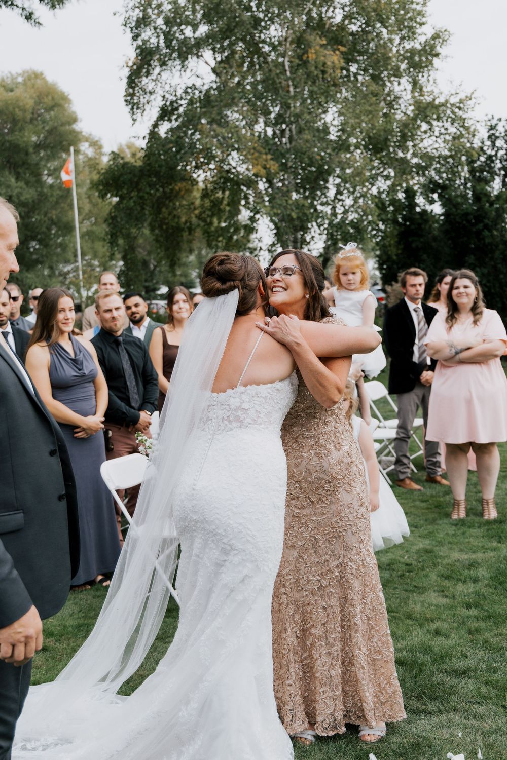 Bride hugging a guest in a gold sequin gown during an outdoor wedding ceremony on a green lawn.