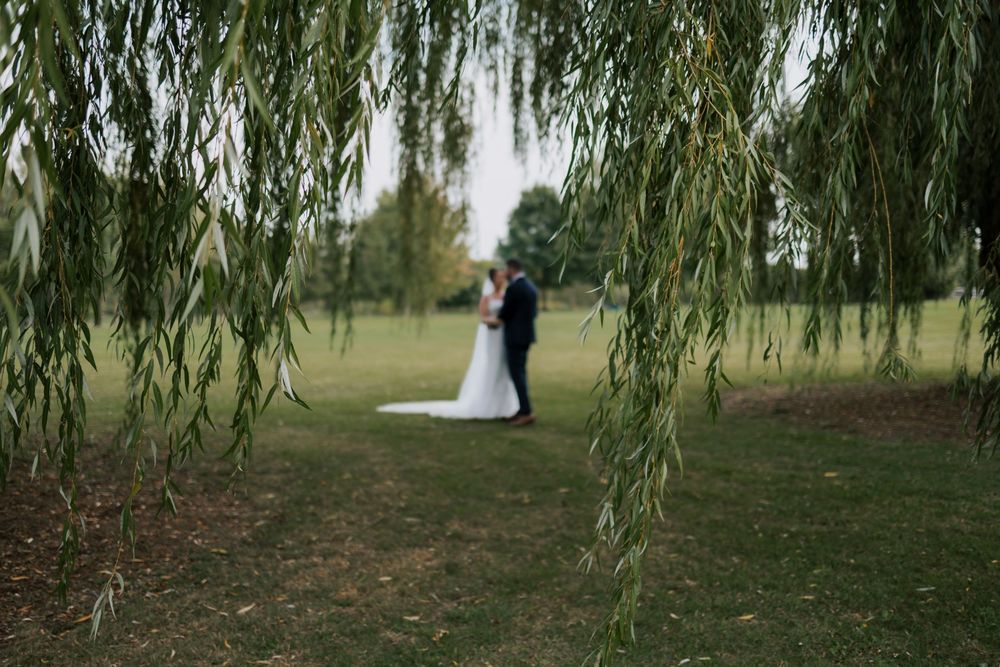 Bride and groom sharing a romantic moment framed by weeping willow branches in a garden.