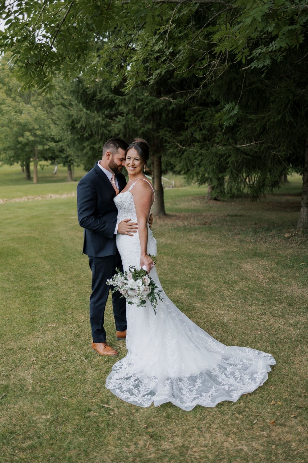 Bride and groom embracing on a green lawn surrounded by tall trees in wedding attire.