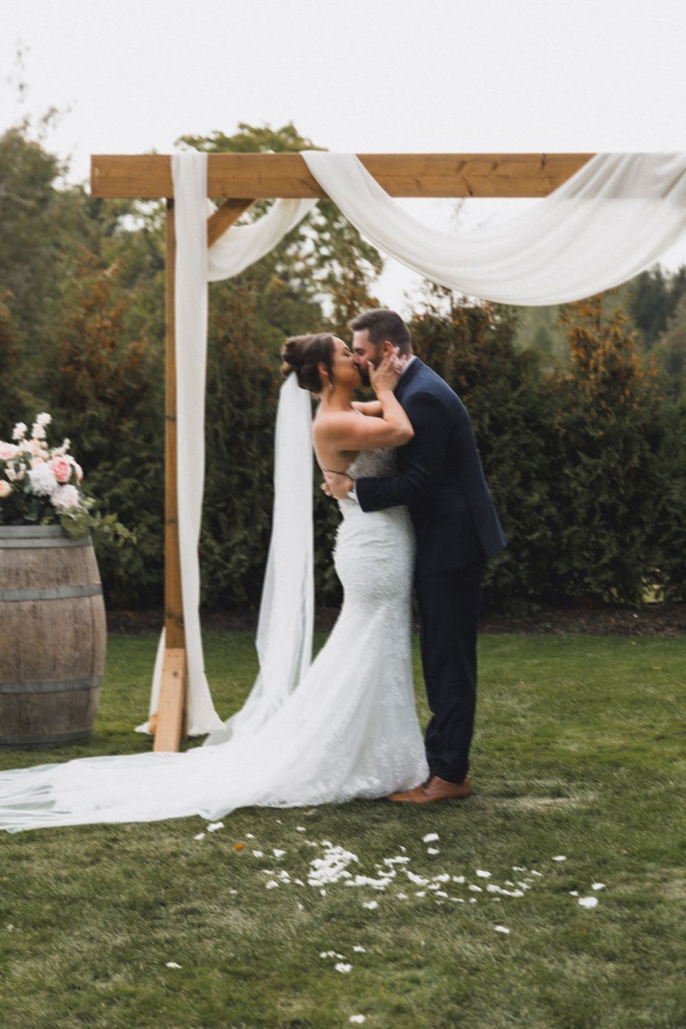Bride and groom share first kiss under rustic wooden arch draped with white fabric at outdoor wedding ceremony.