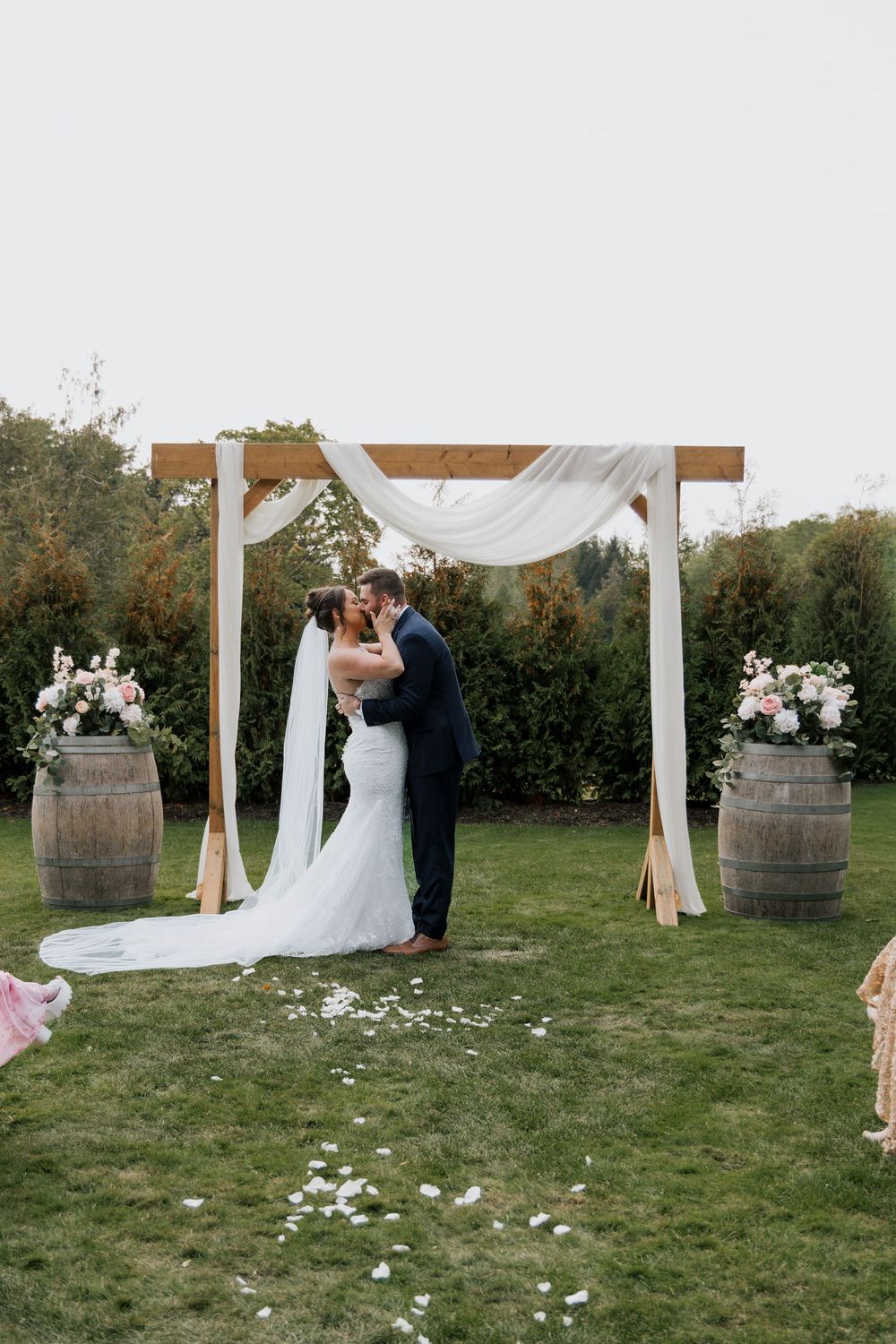 Bride and groom sharing their first kiss under a rustic wooden arch with white draping.