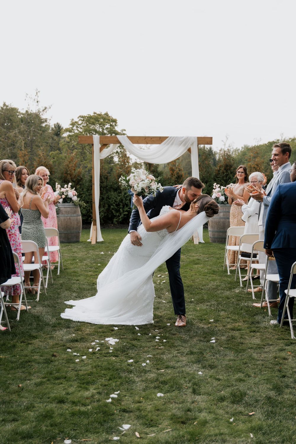 Groom dips bride for romantic kiss while guests applaud along the outdoor wedding ceremony aisle.