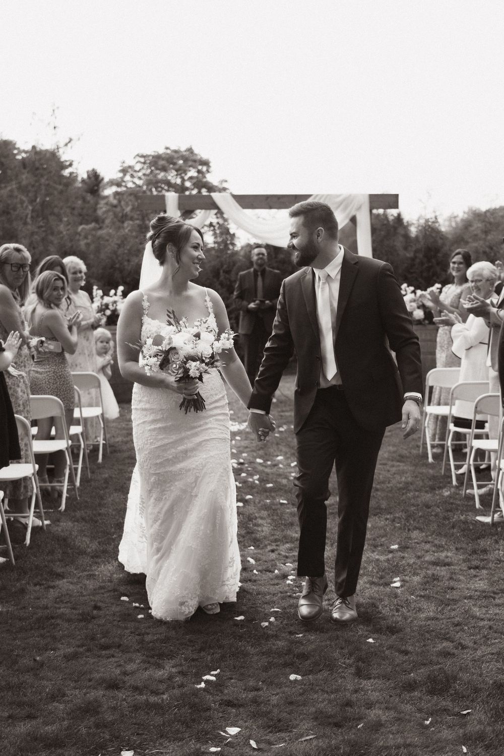 Black and white photo of newlyweds walking hand in hand down the aisle after outdoor wedding ceremony.