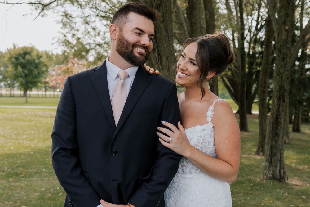 Smiling bride and groom pose together outdoors in wedding attire among trees after their ceremony.