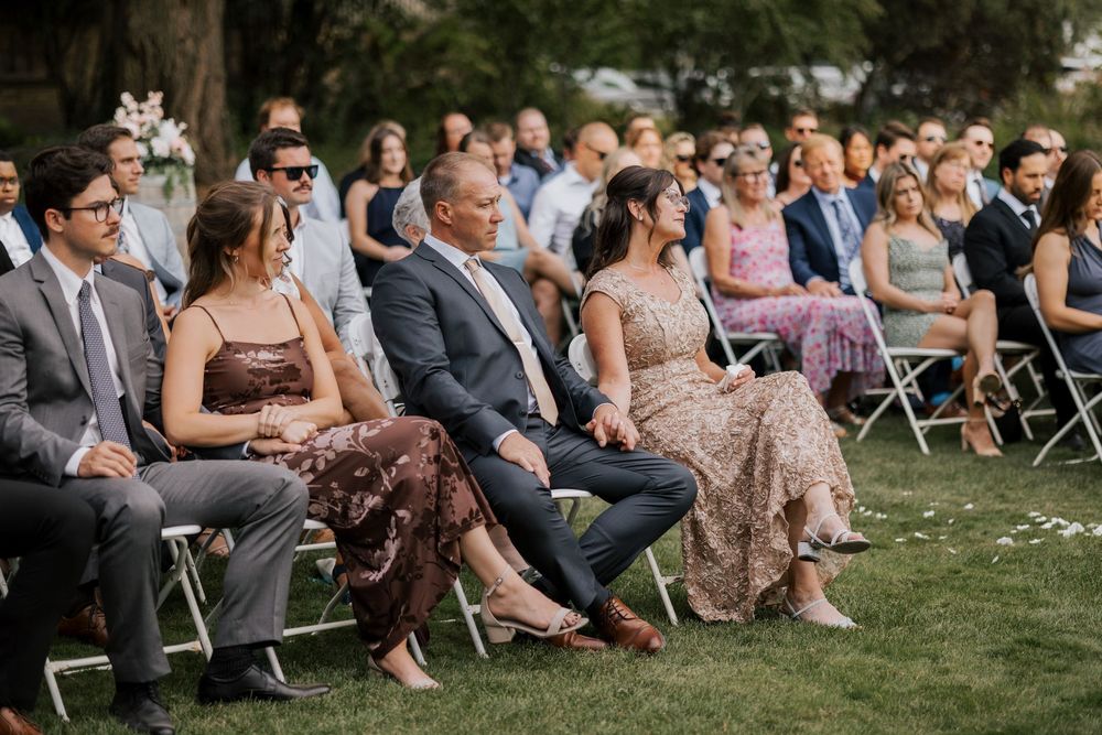 Wedding guests seated in white chairs on grass watching outdoor ceremony on a sunny day.