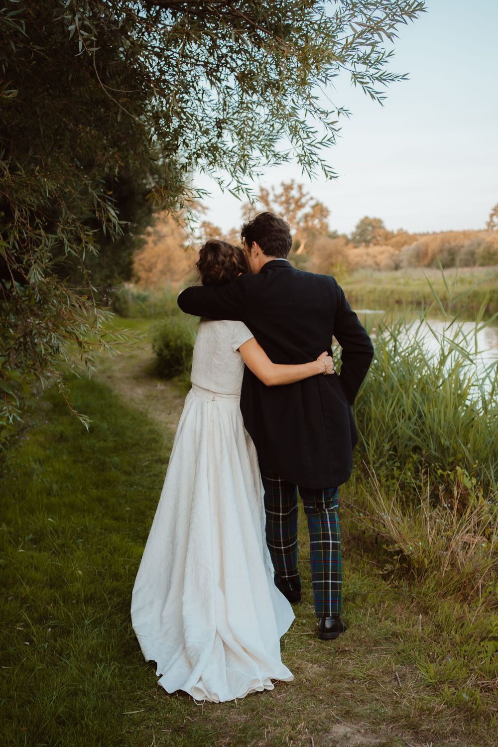 A couple in wedding attire embraces while walking along a riverside path at sunset, with the groom wearing traditional tartan.