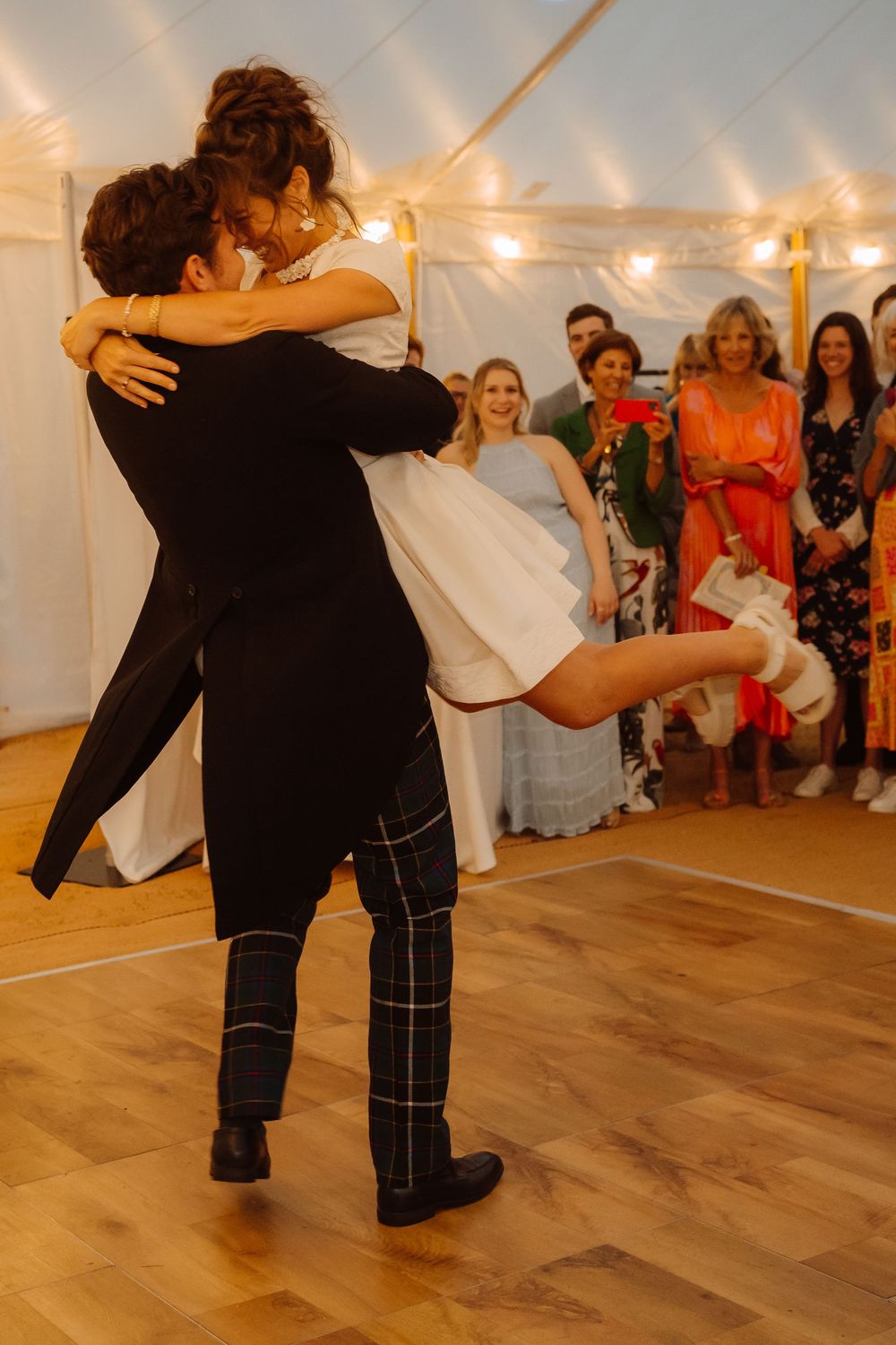 A couple performs a dramatic dance move in a warmly lit room with wooden floors and string lights overhead.